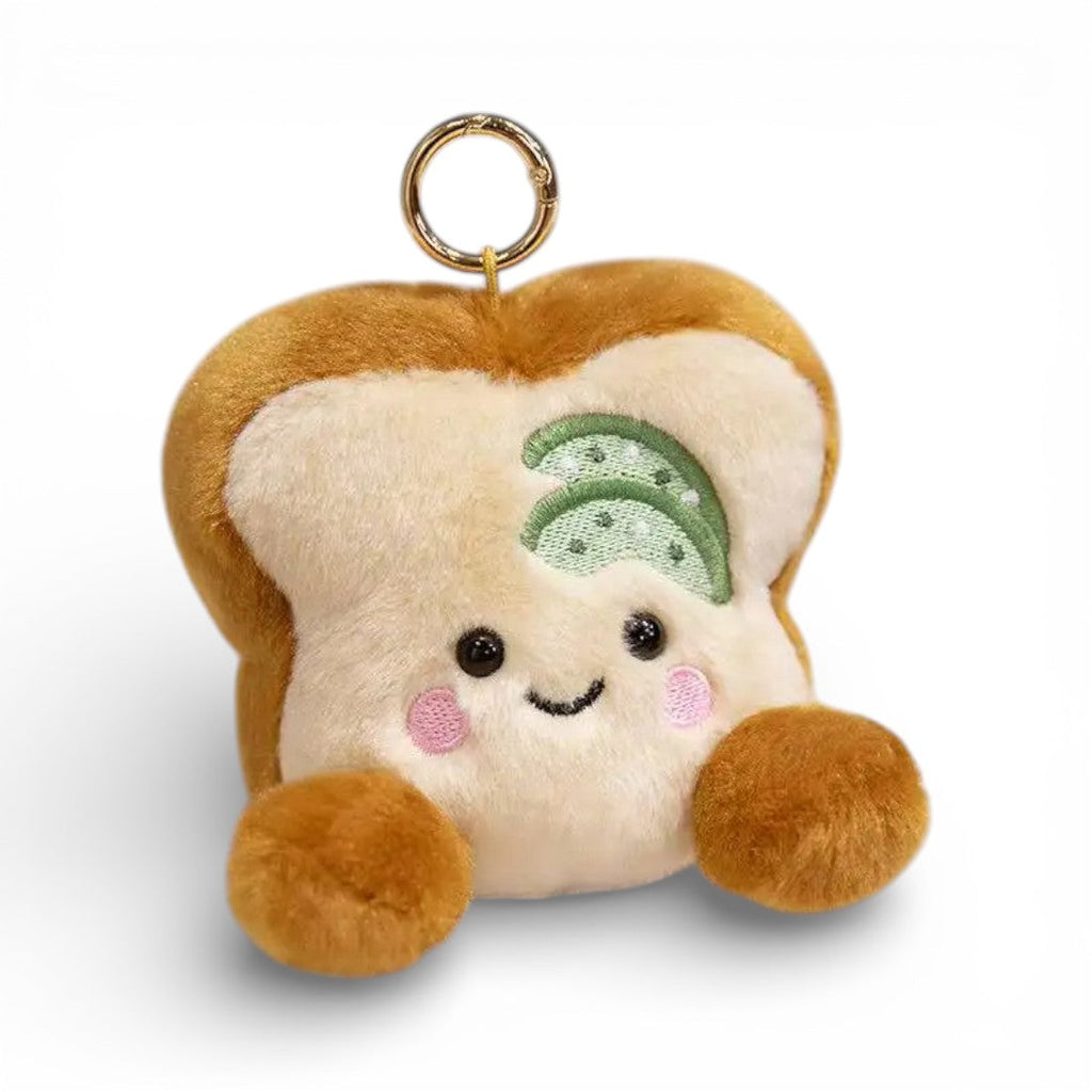 Ein kleiner Plüsch Schlüsselanhänger in Form einer beigen Toastscheibe mit grünem Gurkenbelag obenauf, lächelndes Gesicht mit großen runden Augen und rosa Wangen, kleinen braunen Pfötchen an beiden Seiten, Metallring zur Befestigung, auf weißem Hintergrund.