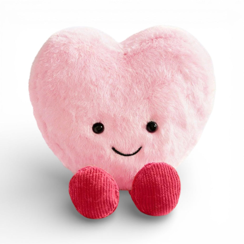 Ein weiches, flauschiges Kuscheltier in Herzform in Rosa, mit einem großen fröhlichen Lächeln, zwei runden glänzenden schwarzen Augen und zwei kleinen roten gestrickten Stoffpfötchen unten, auf einheitlich weißem Hintergrund
