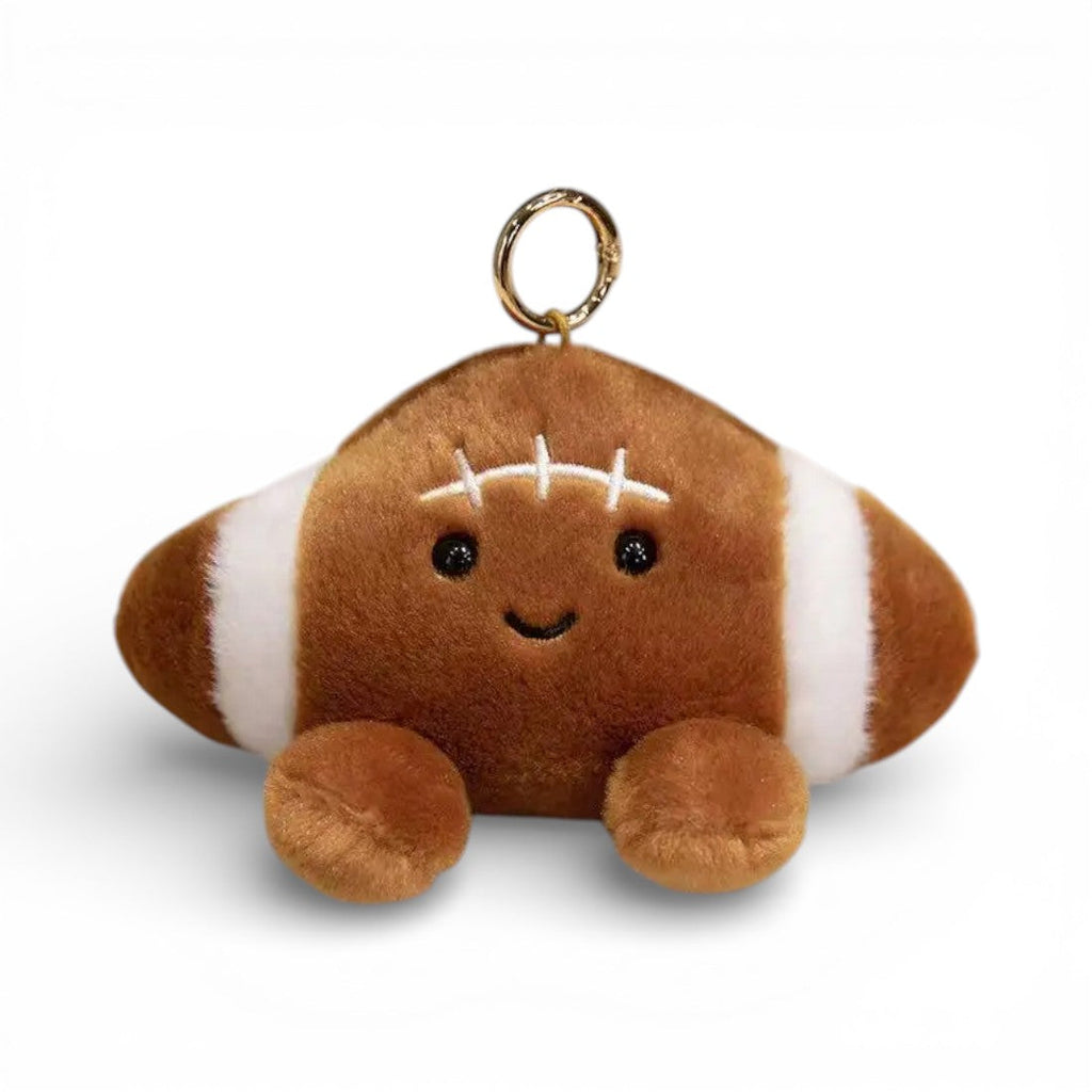 Kawaii-Kuscheltier in Form eines American-Football-Balls, 12 cm, strukturiertes braunes Leder mit weißen Streifen und Nähten, große glänzende schwarze Augen, fröhliches Lächeln, rosa Wangen, zwei pummelige braune Pfötchen unten, goldener Ring obenauf befestigt, auf weißem Hintergrund.